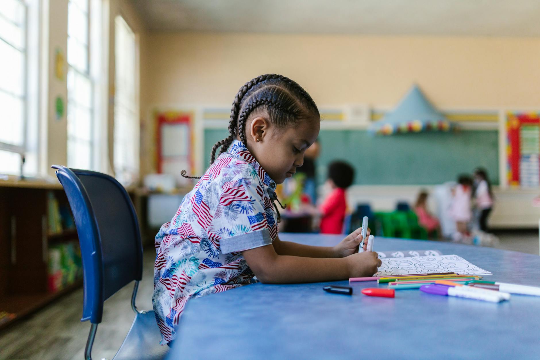 A young child with braids engaged in coloring at a classroom desk surrounded by school supplies.