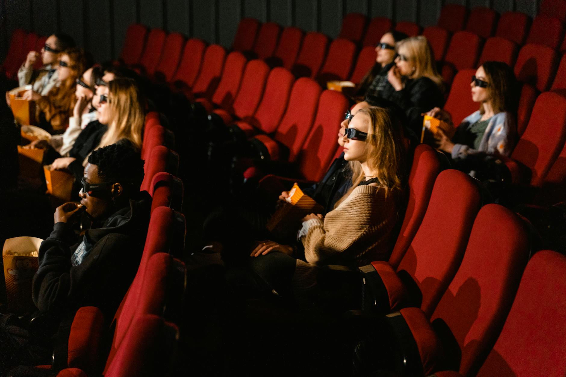 People wearing 3D glasses and eating popcorn in a cinema with red chairs.