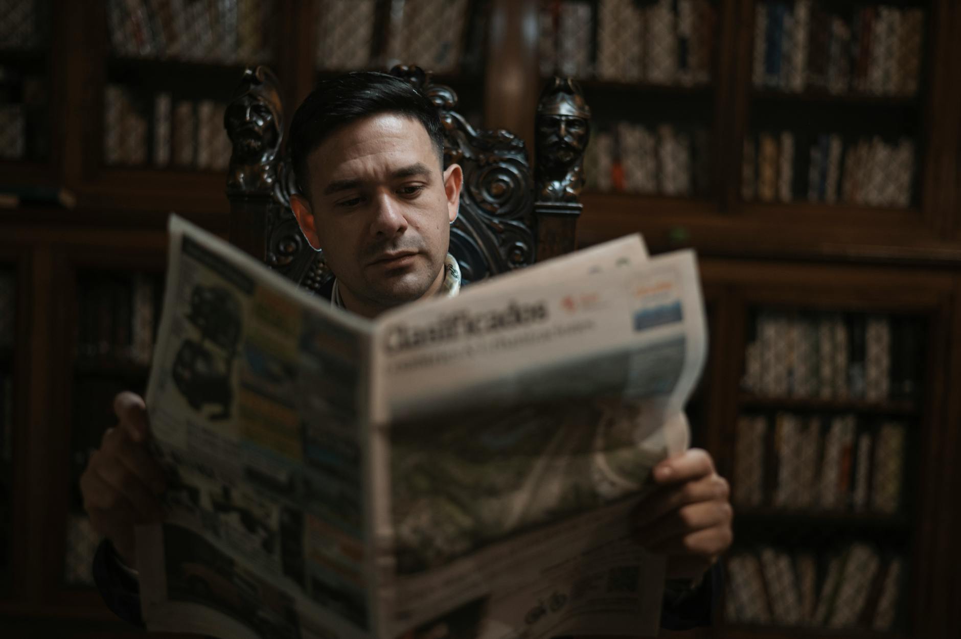A man in an ornate chair reads a newspaper in a sophisticated library setting.