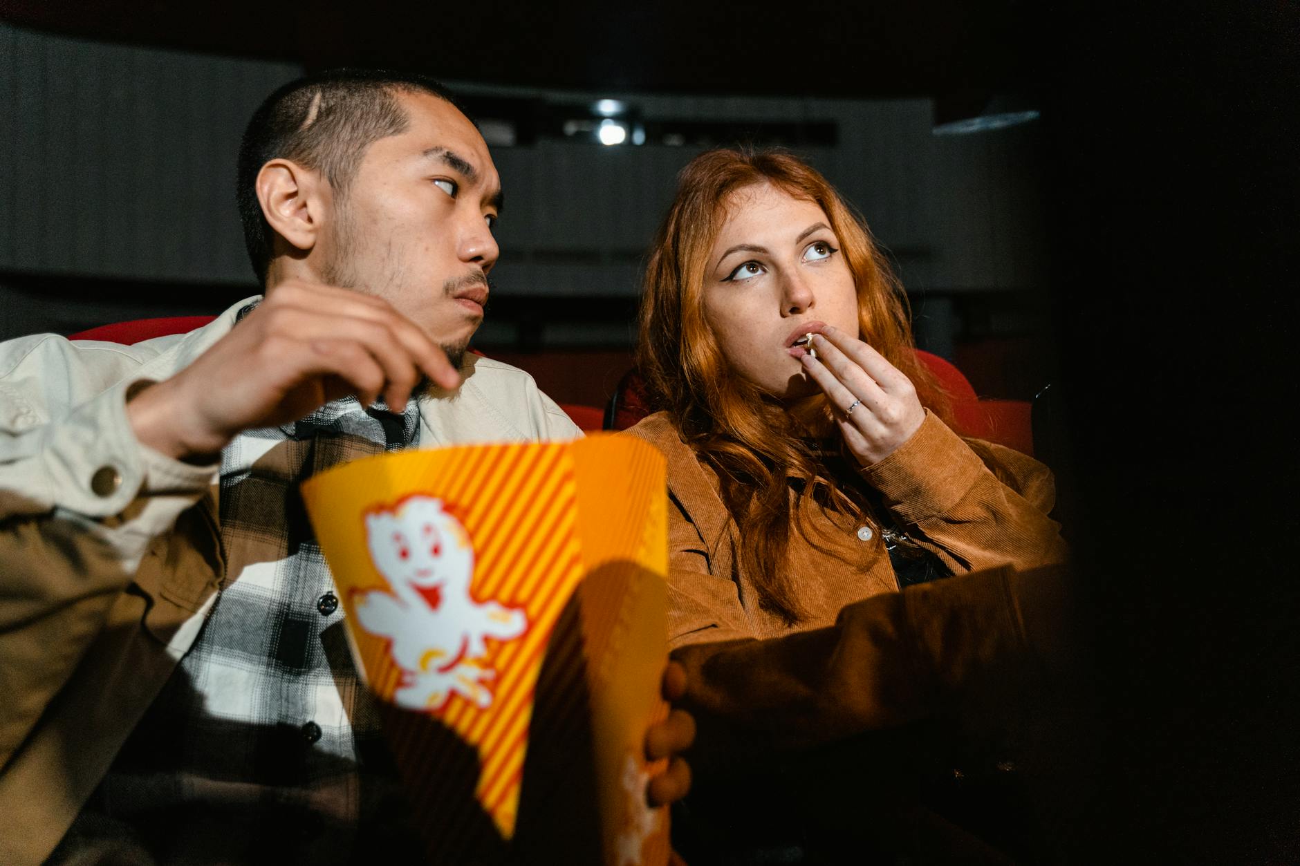 An Asian man and Caucasian woman enjoying popcorn together in a cozy movie theater.
