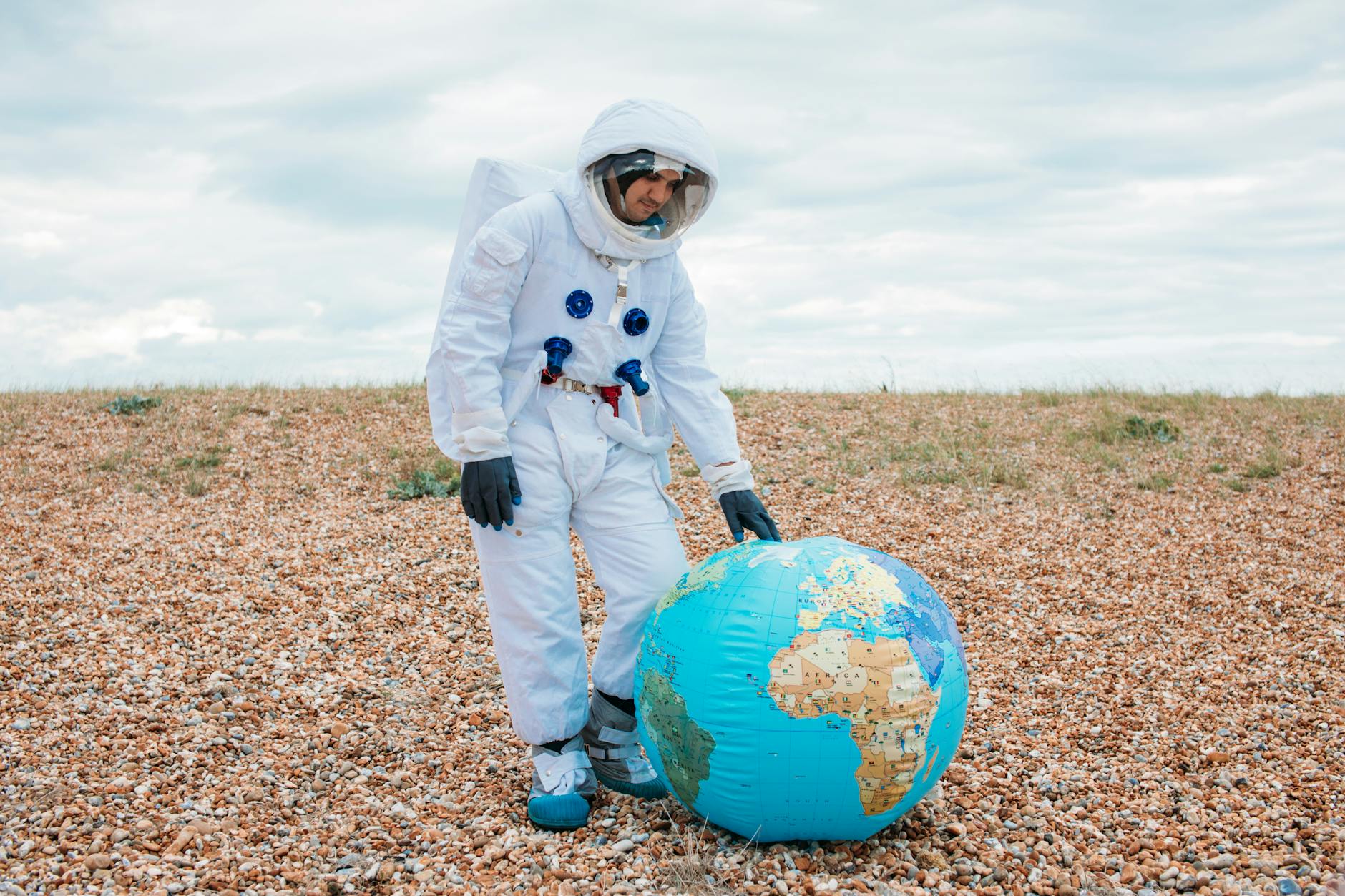 An astronaut in a white space suit on a pebble beach, exploring an inflatable globe under a cloudy sky.