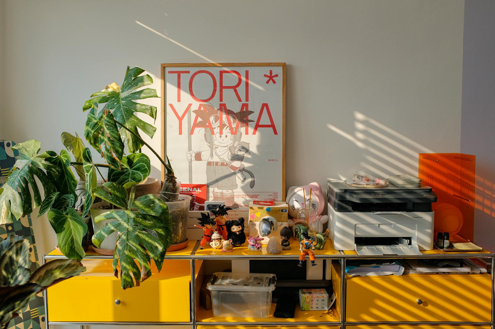 A vibrant workspace features anime decor, houseplants, and a printer bathed in natural light.