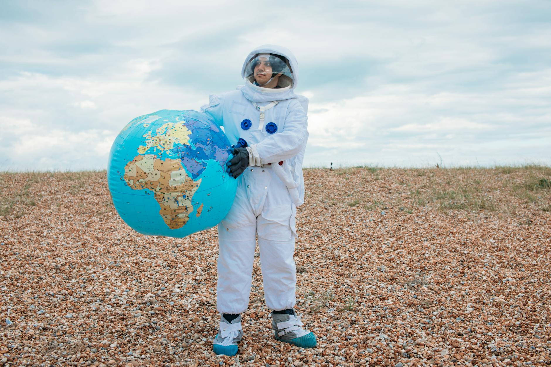 Astronaut in a space suit holding a large Earth globe outdoors on a rocky terrain.