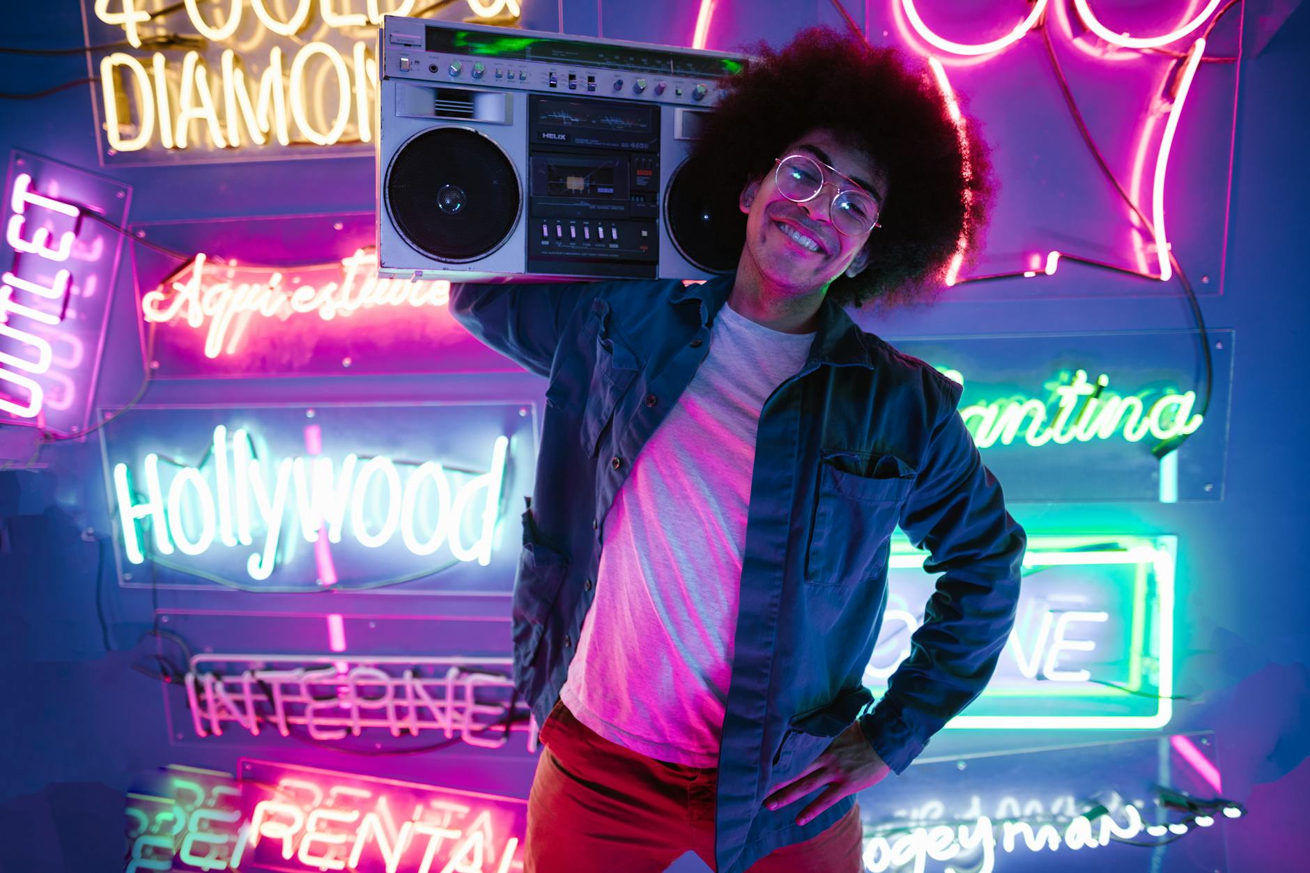 A young man with afro hair smiling, holding a vintage boombox in a vibrant neon-lit room.