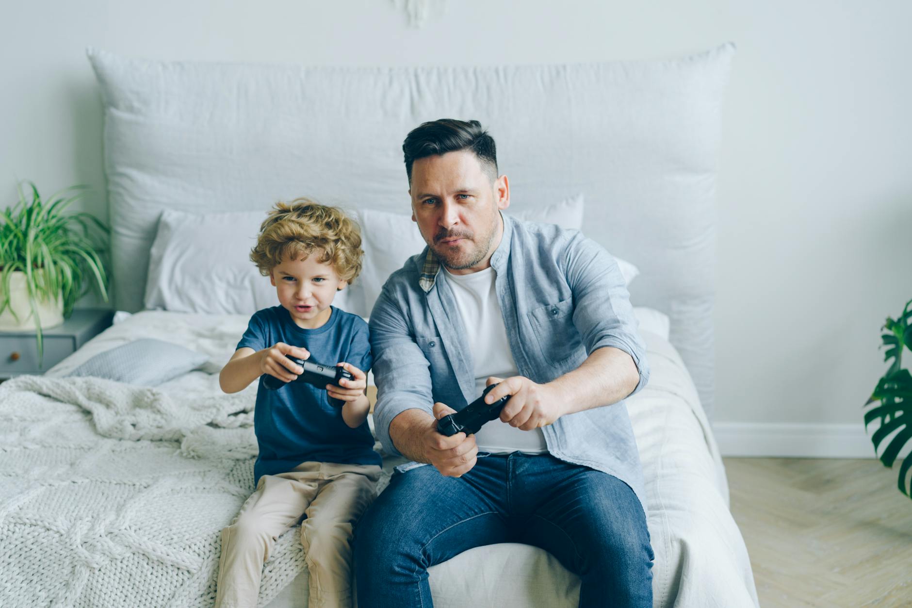 Father and son joyfully playing video games while sitting on a bed, capturing a moment of family fun.