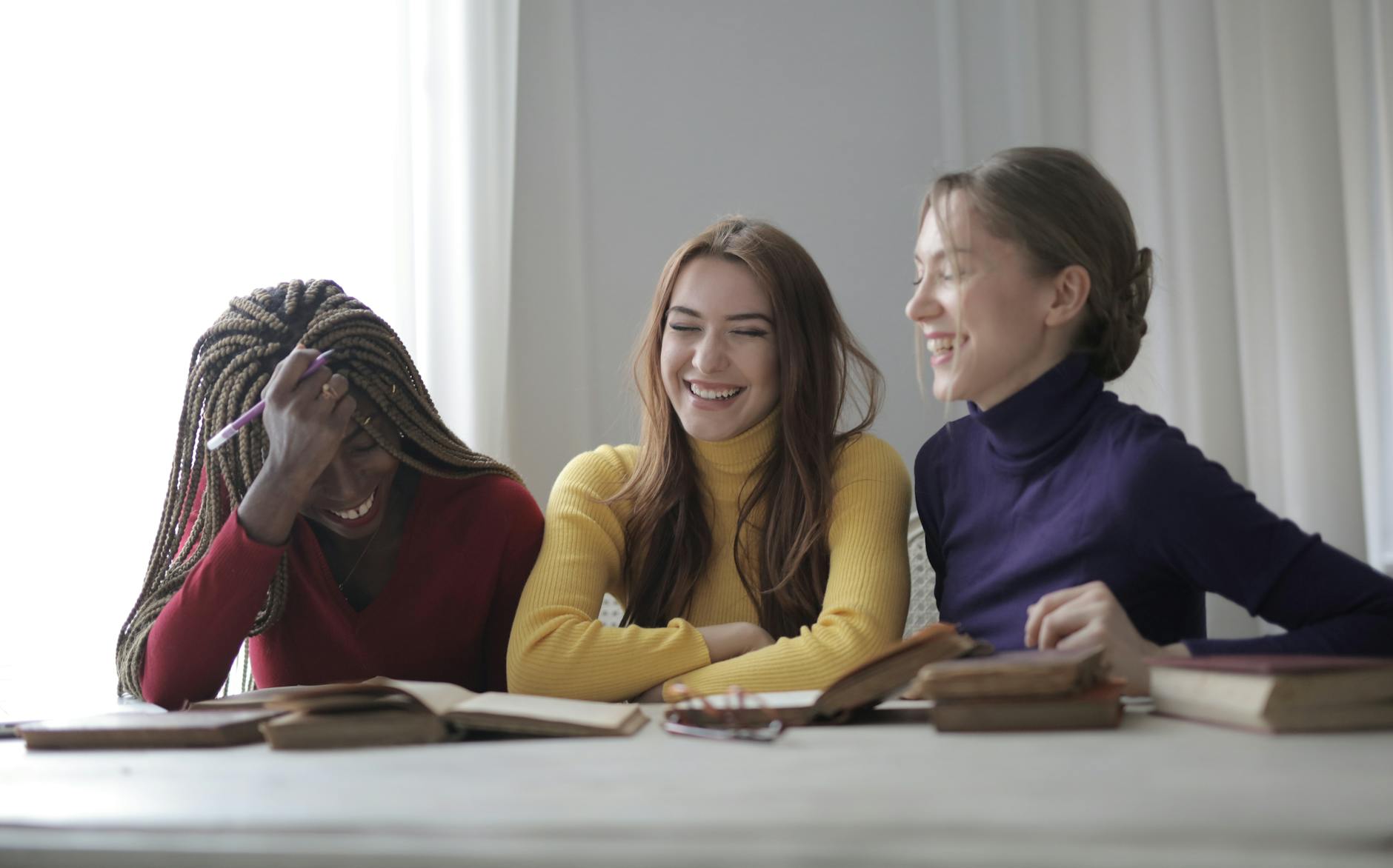 Cheerful young multiracial women in colorful casual clothes laughing while sitting together at table with books in modern light room