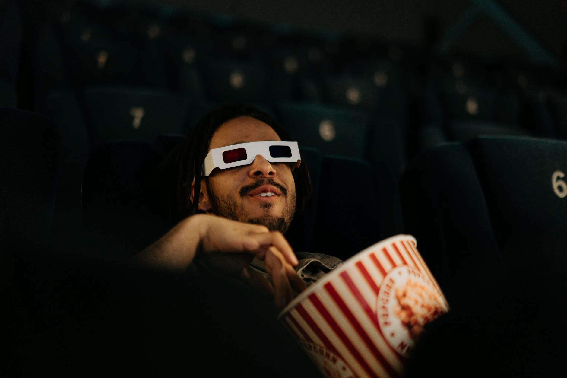 A man wearing 3D glasses enjoys popcorn while watching a movie in a cinema.