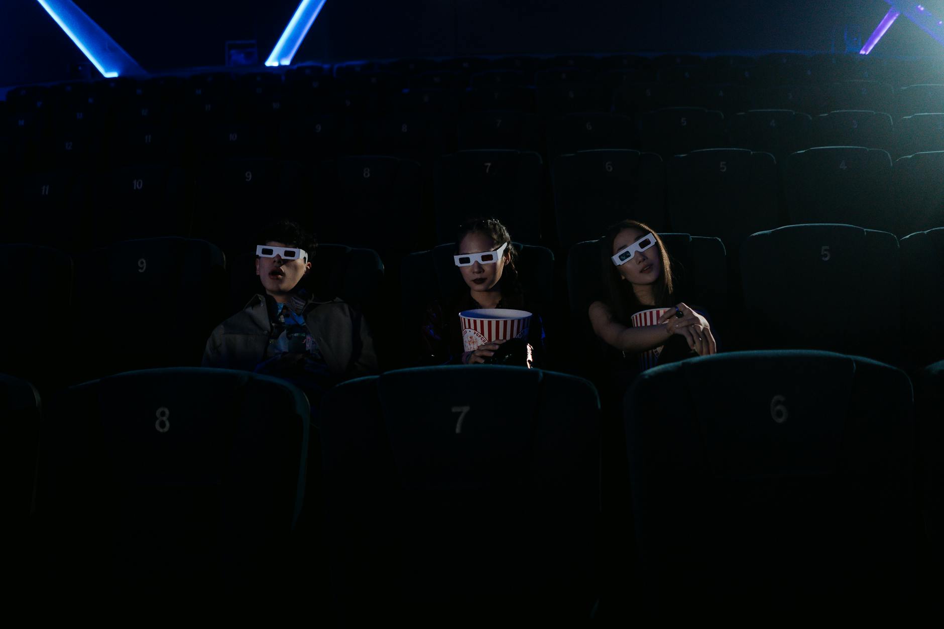 Three friends enjoying a movie with popcorn and 3D glasses in a dimly lit cinema.