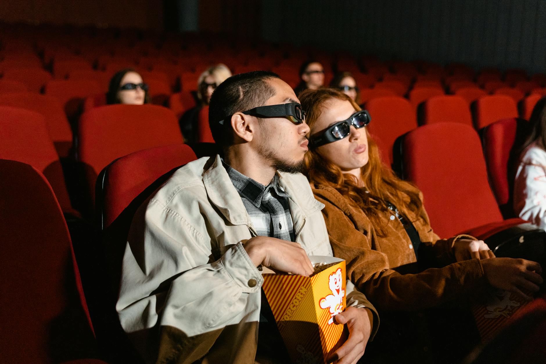 Couple wearing 3D glasses watching a movie in a cinema, enjoying popcorn.