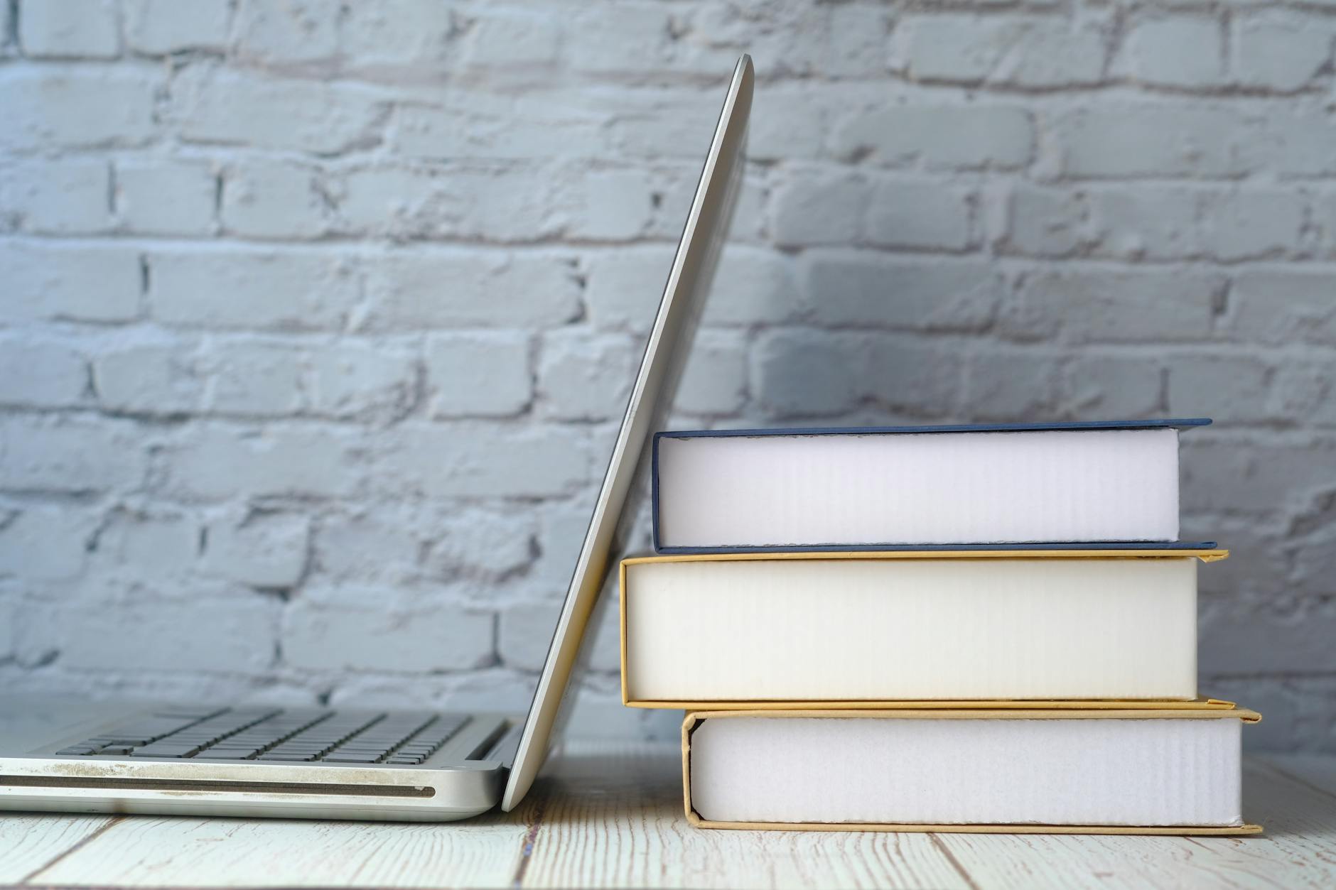 A stack of books next to an open laptop on a wooden desk against a brick wall.