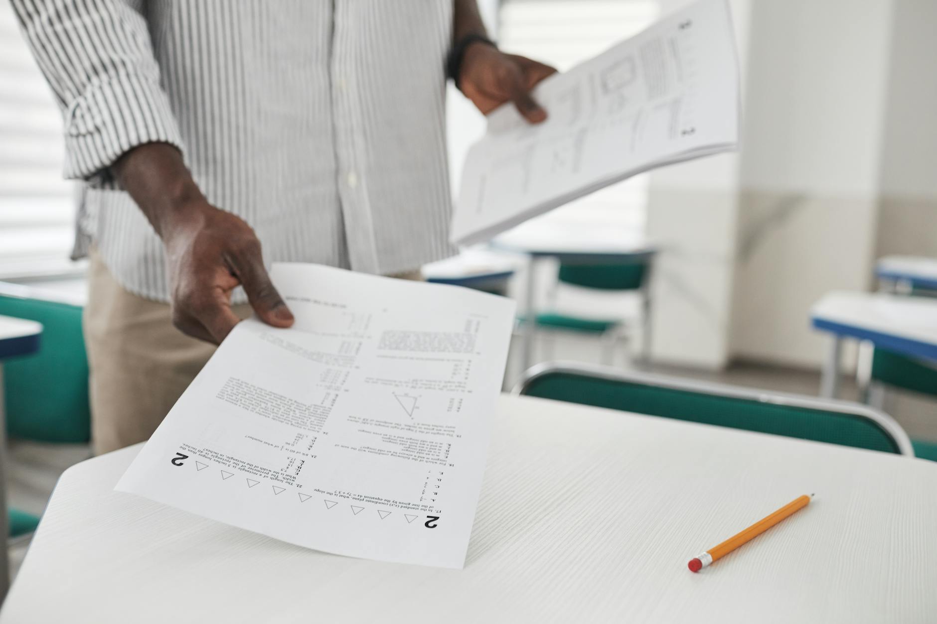 Close-up of a person arranging examination papers on a desk in a classroom.