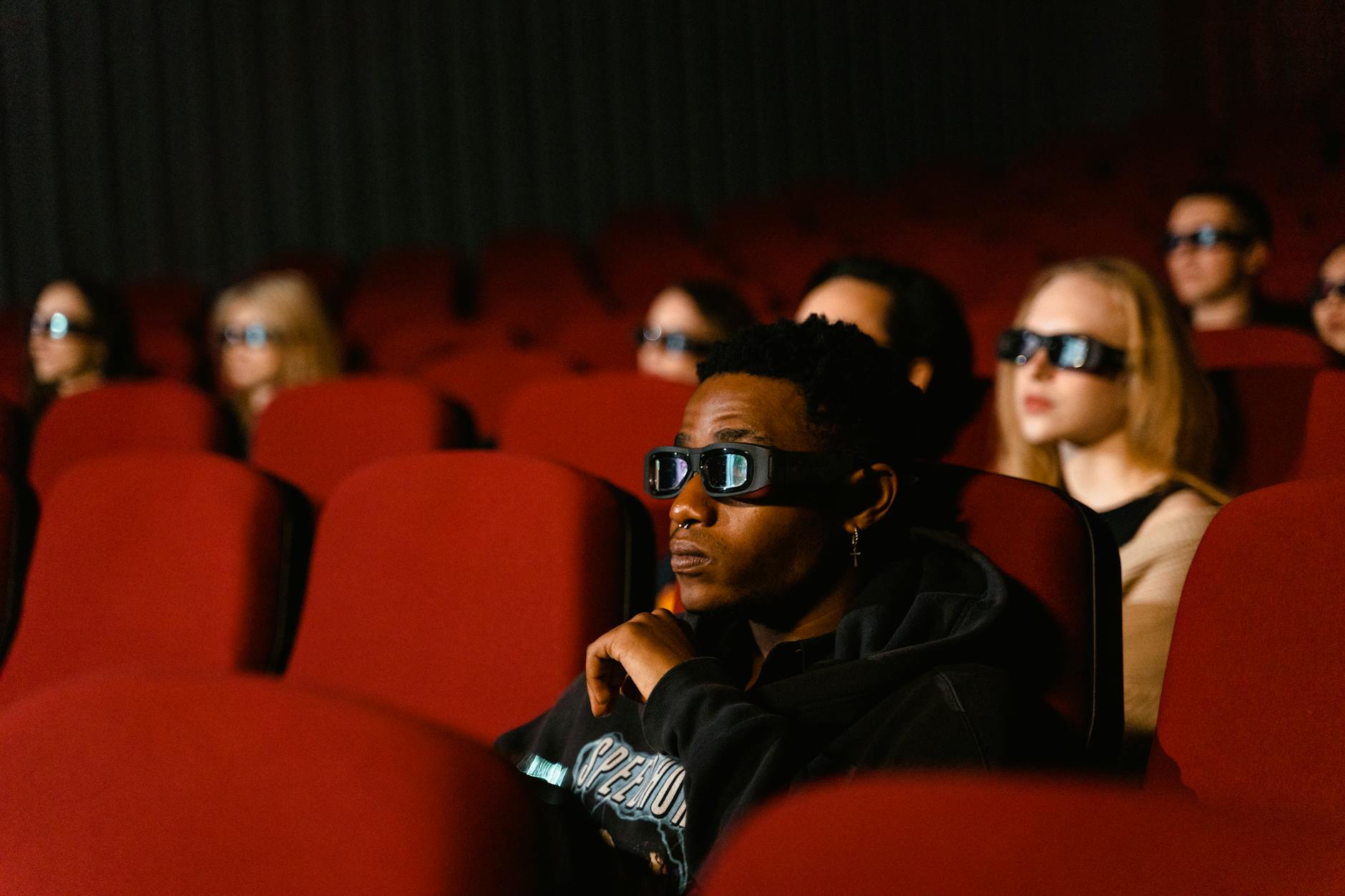 A diverse group of people wearing 3D glasses enjoying a movie in a cinema with red seats.