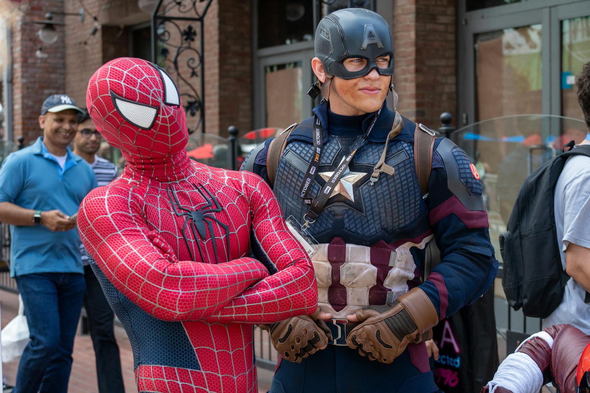 Spider-Man and Captain America cosplayers pose in a lively street scene with onlookers.