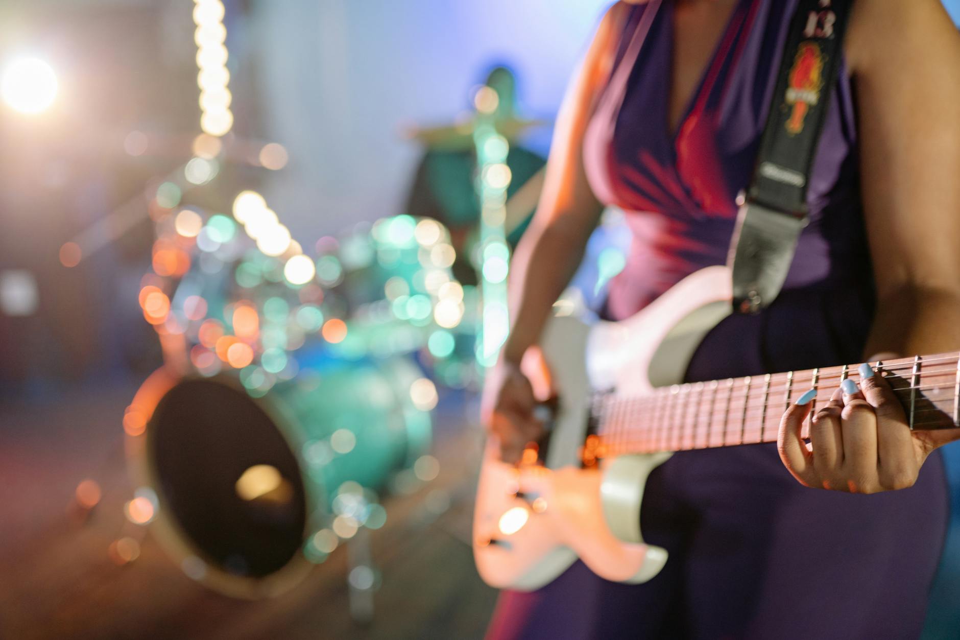 Close-up of a female guitarist playing on stage with drum set in background.