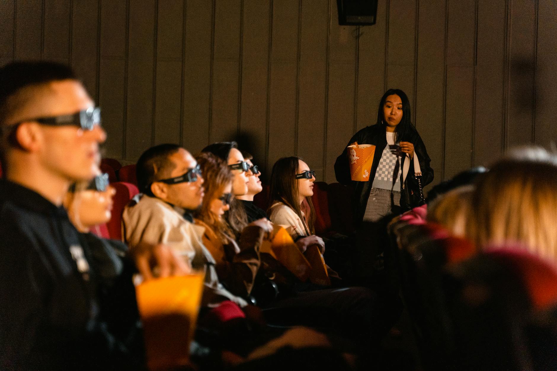 A group of diverse young adults watching a 3D movie indoors, enjoying popcorn.