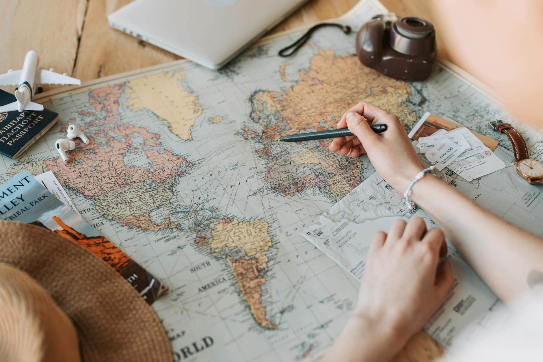 Hands marking destinations on a world map surrounded by travel essentials like passport and camera.