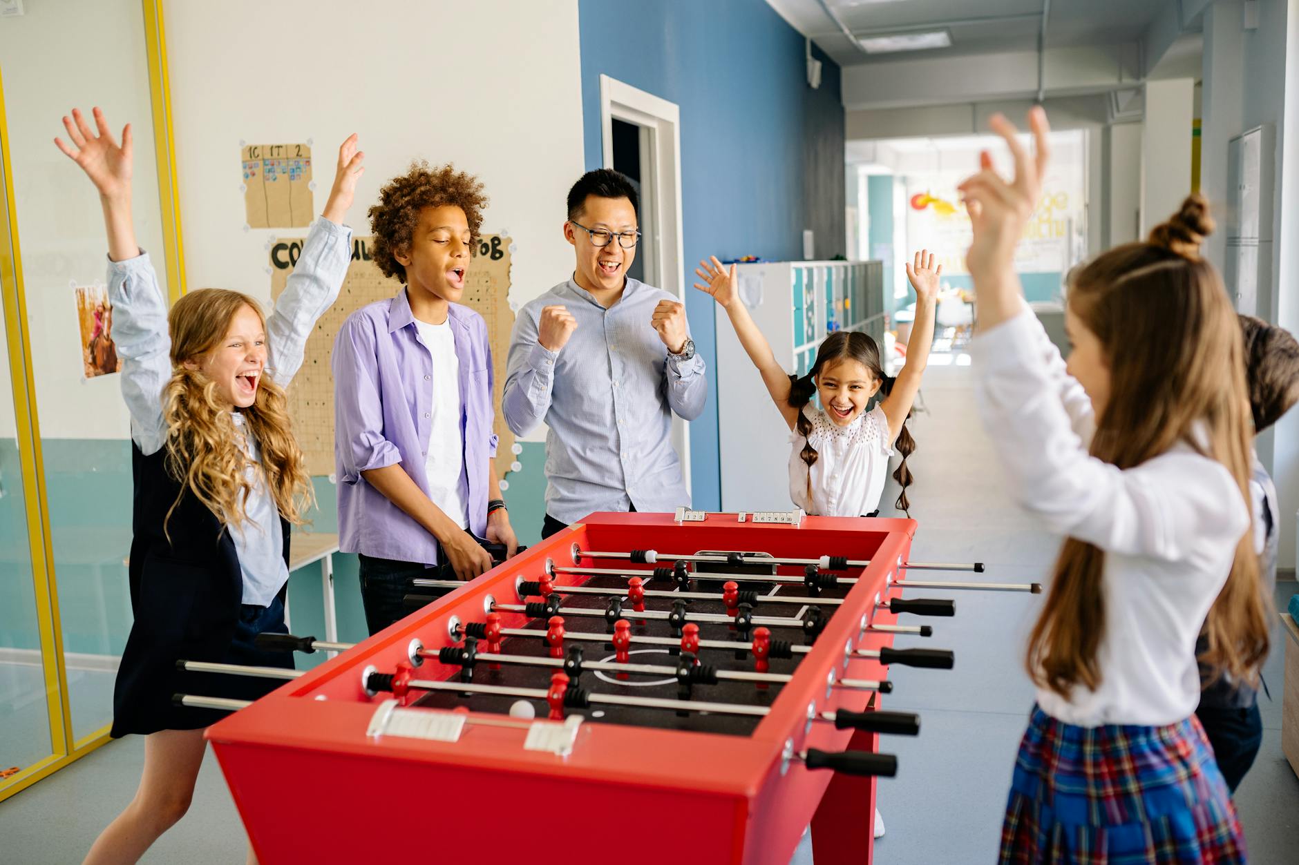 A diverse group of students and a teacher playing foosball indoors, celebrating a win.