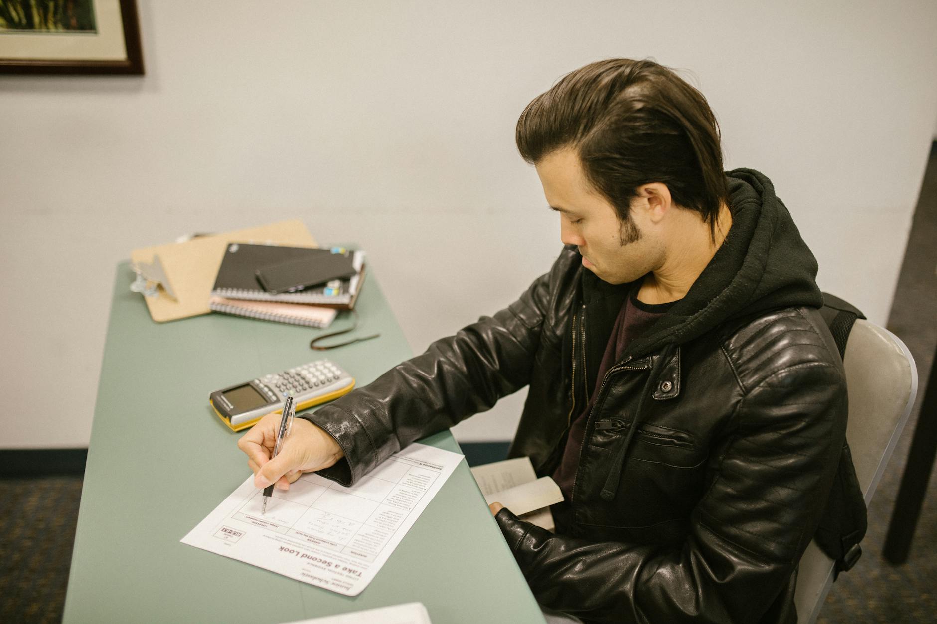 Man in leather jacket writing test answers in college classroom setting.