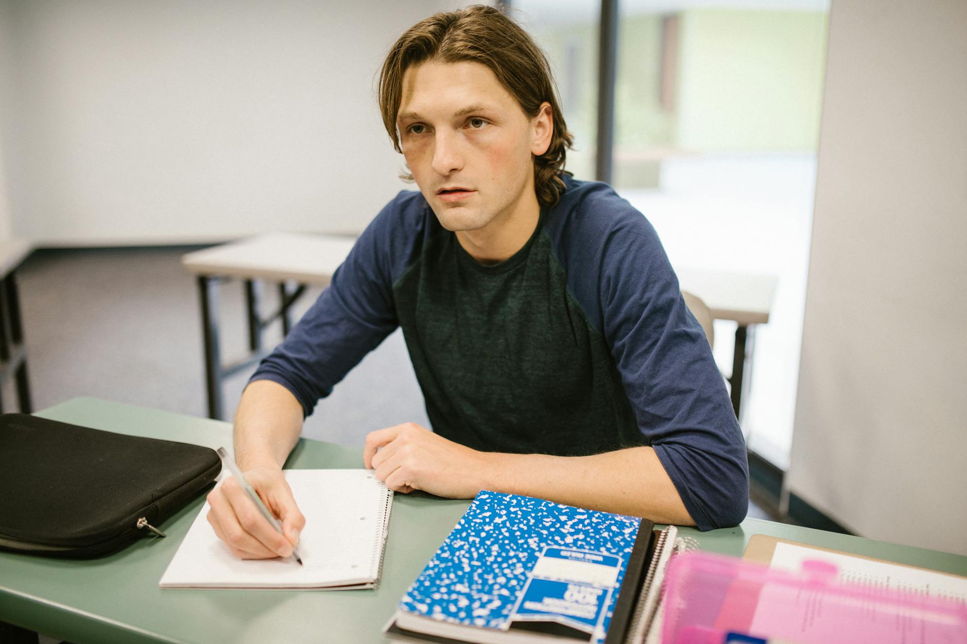 Young man studying in a classroom, focused on writing notes with a pen.