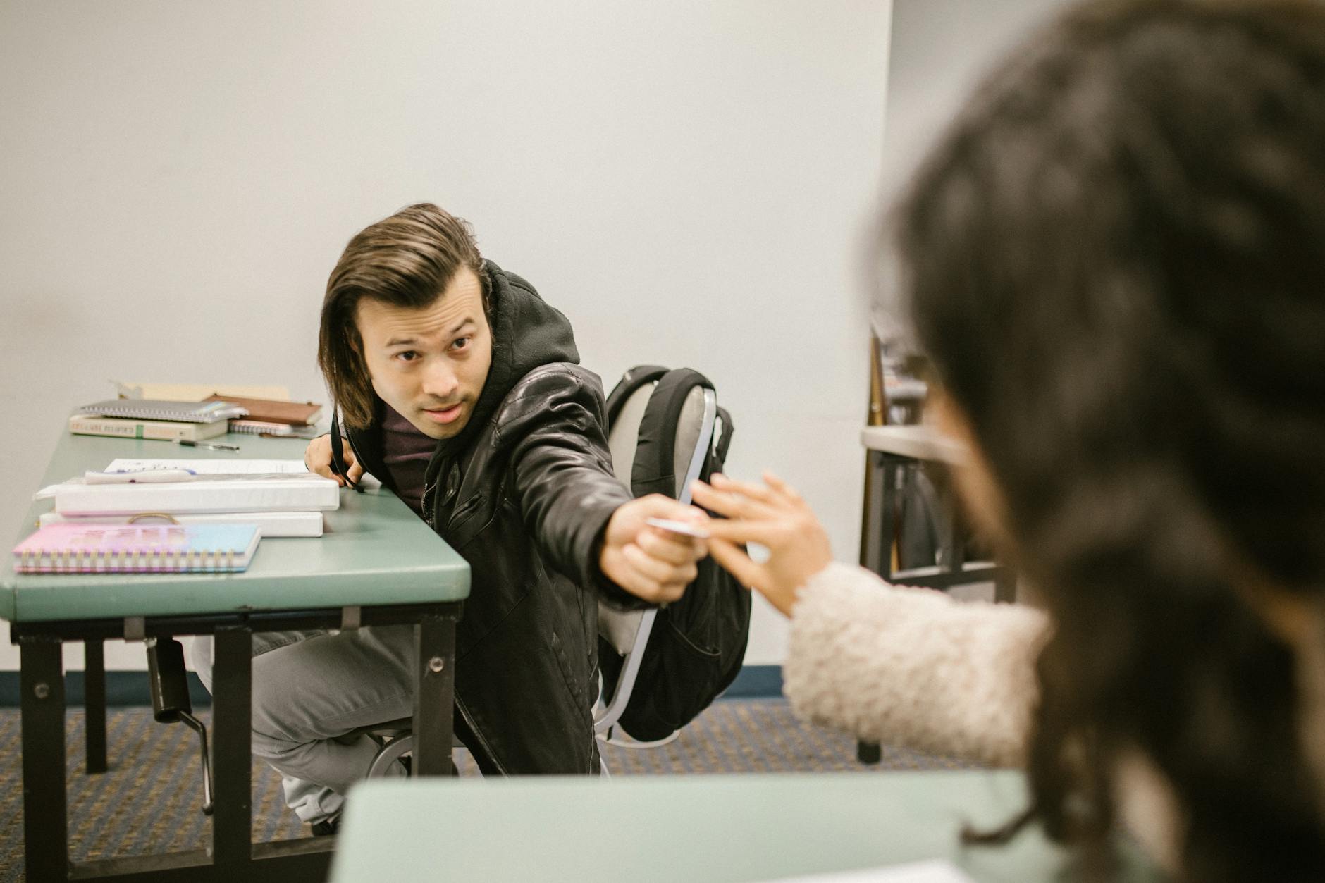 Male student discreetly passing a note to a classmate during an exam in a college classroom.